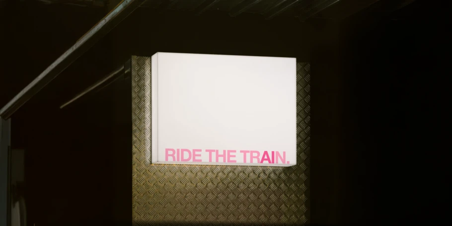 Image of a rectangular lit sign set on a sheet of corrugated metal. The light is bright white and the text is in all caps in pink with the letters "AI" subtly set apart in a darker pink.. TEXT: "RIDE THE TRAIN."